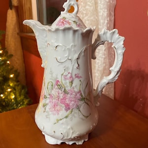 May include: White porcelain coffee pot with a lid, featuring pink floral designs and gold trim. The pot has an ornate handle and spout, with a decorative finial on the lid. The overall design is vintage and elegant.