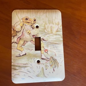 May include: A decorative light switch cover featuring a whimsical illustration of a frog fishing in a pond. The frog is dressed in a coat and pants, with a fishing rod in hand. The color palette includes shades of green, beige, and brown.
