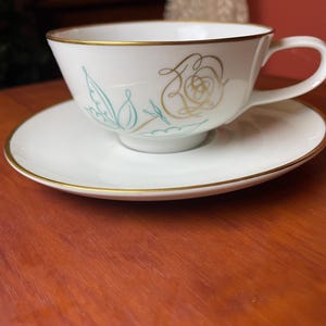 May include: White teacup and saucer set with gold trim. The cup features a floral design in gold and teal. The saucer and cup have a gold trim. The set is on a wooden surface.