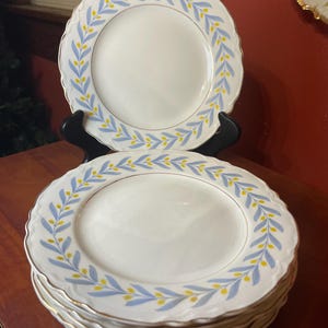 May include: A stack of white ceramic plates with a blue and yellow leaf design around the rim. The plates have a scalloped edge and a gold trim. The plates are displayed on a wooden surface.