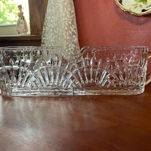 May include: A clear, rectangular crystal serving dish with two handles. The dish features a cut glass design with a repeating pattern of diamond shapes and fan-like details. The dish is likely used for serving food or displaying items.