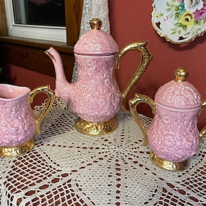 May include: A pink ceramic tea set with a floral pattern, featuring a teapot, creamer, and sugar bowl. The set has gold-colored handles and bases, and a gold finial on the teapot and sugar bowl.