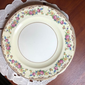 May include: A vintage cream-colored dinner plate with a gold rim and a floral pattern featuring pink, yellow, and blue flowers.