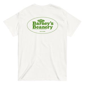 Può includere: T-shirt bianco con un design ovale verde. Il design presenta il testo "Barney's Beanery" in verde, con le parole "Pool & Games" in basso. La maglietta è realizzata in un materiale morbido.