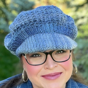 May include: A woman wearing a blue knitted hat with a brim and black framed glasses. She is wearing a blue denim jacket.