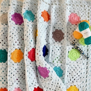 May include: A light blue crochet blanket with colorful granny squares featuring a circle design. The blanket is folded in thirds and yarn balls are visible in the background.