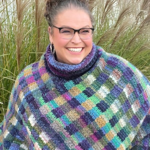 May include: A woman wearing a colorful, crocheted poncho with a high neck. The poncho has a checkered pattern in shades of blue, green, purple, and brown.