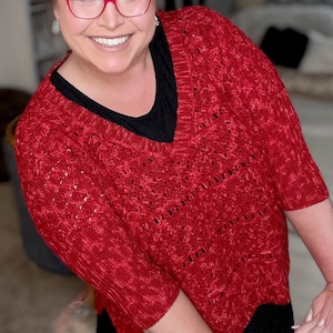May include: A woman wearing a red knitted sweater with a black v-neck undershirt and a black skirt. She is smiling and wearing red-rimmed glasses.