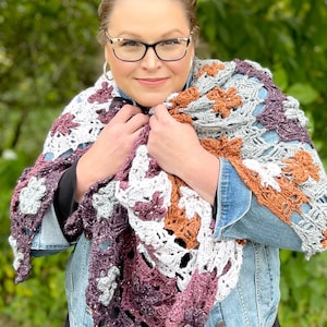 May include: A woman wearing a denim jacket and a crocheted shawl with a multi-colored floral pattern. The shawl is made with different shades of purple, white, gray, and orange yarn.