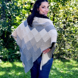 May include: A woman wearing a crocheted shawl with a chevron pattern in shades of gray and white. The shawl is draped over her shoulders and tied at the waist. She is standing in a grassy area with trees in the background.