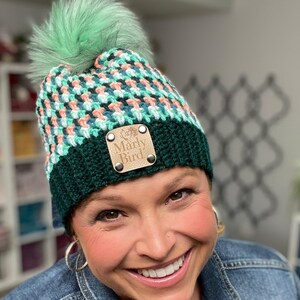 May include: A handmade crochet beanie with a green and peach pattern, topped with a fluffy green pom-pom. The hat features a dark green band and a leather tag that reads "Marly Bird".