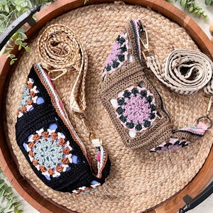 May include: Two crochet crossbody bags with adjustable straps rest on a round woven tray. One bag is black with a colorful floral design, the other is brown with a purple floral design. The straps are beige and cream.