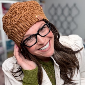 May include: A textured, rust-colored crochet beret featuring a ribbed brim and a small, rectangular brown leather tag with the text Marly Bird embossed in gold. The hat has a complex, repeating stitch pattern throughout the crown.