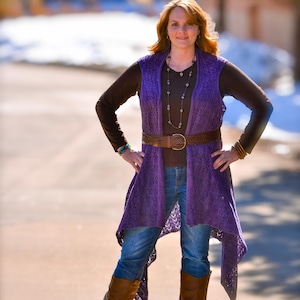 May include: A woman wearing a purple open-front vest over a brown long-sleeve shirt and blue jeans. She is also wearing a brown belt, brown boots, and a necklace. The vest has a lace-like design.