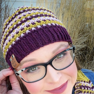 May include: A knitted beanie in shades of purple, yellow, and pink, with a matching scarf. The beanie has a leather tag that reads "Marly Bird". The glasses have black frames with a white and gray pattern.