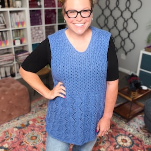 May include: A blue knitted vest with a V-neck. The vest has a detailed openwork pattern. The person is wearing the vest over a black top and blue jeans. The background includes a bookshelf and a patterned rug.