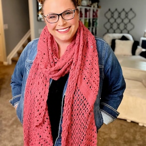 Może przedstawiać: Różowy szydełkowy szal z delikatnym ażurowym wzorem. Szal jest noszony na dżinsowej kurtce. Kolor szala to stonowany róż, a ażurowy wzór dodaje tekstury. Ręcznie robiony dodatek.