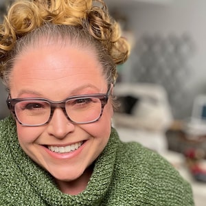 May include: A woman wearing a green knitted scarf and a denim jacket. She is smiling and wearing glasses.