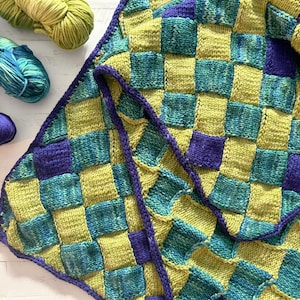 May include: A handmade knitted blanket with a checkerboard pattern in shades of green, blue, and purple. The blanket is made of soft yarn and has a textured appearance. Several skeins of yarn in matching colors are visible.