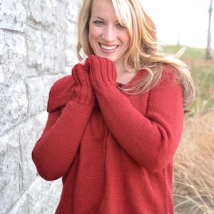 May include: A red knit sweater with a wide collar and long sleeves. The sweater is a solid color and appears to be made of a soft, textured material. The person is wearing blue jeans.
