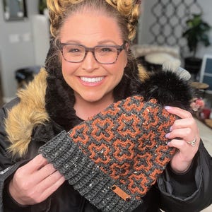 May include: A grey and orange crocheted beanie with a black pom pom. The beanie has a brown leather patch on the side.