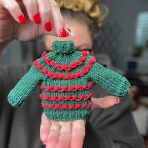 May include: A green knitted sweater ornament with red decorative trim. The miniature sweater has long sleeves and a turtleneck collar. The ornament is held up by a silver hook.