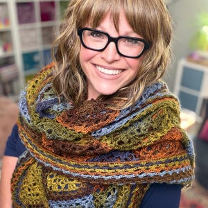 May include: A crochet shawl featuring a multi-colored pattern of olive green, deep brown, and muted blue yarn. The accessory is constructed with an intricate, open-work stitch design that creates a textured, layered appearance across the fabric.
