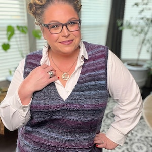 May include: A woman wearing a white shirt and a purple and blue striped knitted vest. She is wearing glasses and a necklace with a circular pendant.