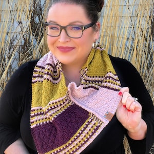 May include: A woman wearing a black top and a colorful knitted scarf with stripes of pink, purple, and yellow. The scarf is folded in a triangular shape and has a brown leather tag.