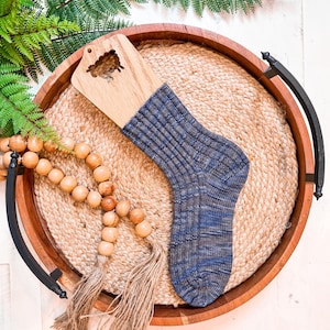 A blue knit sock on a wooden sock blocker, displayed on a round woven mat in a wooden tray with handles—perfect as a handmade gift—alongside a strand of wooden beads with tassels and a green fern.