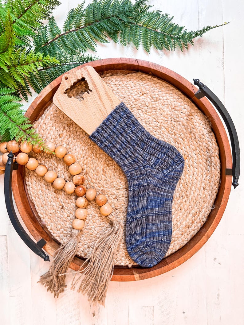 A blue knit sock on a wooden sock blocker, displayed on a round woven mat in a wooden tray with handles—perfect as a handmade gift—alongside a strand of wooden beads with tassels and a green fern.