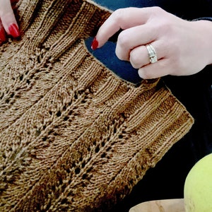 A person with red nail polish is Holding a knitting a brown textured market bag, The image shows their hands actively holding the hand hole on the bag. A bowl with a green pear is partially visible