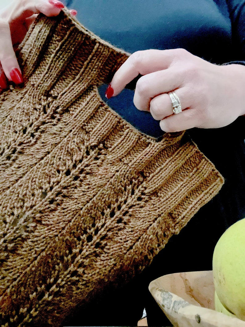 A person with red nail polish is Holding a knitting a brown textured market bag, The image shows their hands actively holding the hand hole on the bag. A bowl with a green pear is partially visible