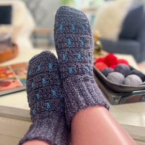 May include: A pair of gray crocheted socks with blue accents. The socks are worn by a person sitting on a couch.