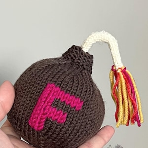 May include: A brown knitted bomb-shaped toy with a pink letter "F" on the front. The bomb has a white knitted fuse and a tassel of yellow, red, and pink yarn. The toy is held in a hand.