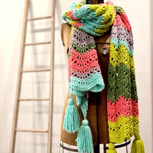 A vividly colored Calor Crochet Wrap by Marly Bird draped over a mannequin, showcasing a wave crochet pattern and bright tassels.
