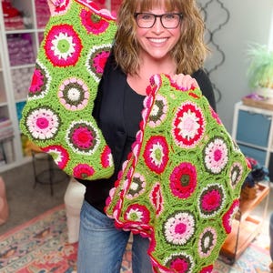 May include: A crochet Christmas stocking and a matching rectangular blanket featuring a vibrant floral motif. The items are constructed from joined circular granny squares in shades of lime green, hot pink, light pink, white, and dark olive green.