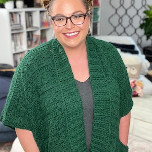 May include: A woman wearing a dark green cardigan with pockets. The cardigan has a cable knit pattern and is open in the front. She is wearing a gray t-shirt underneath.