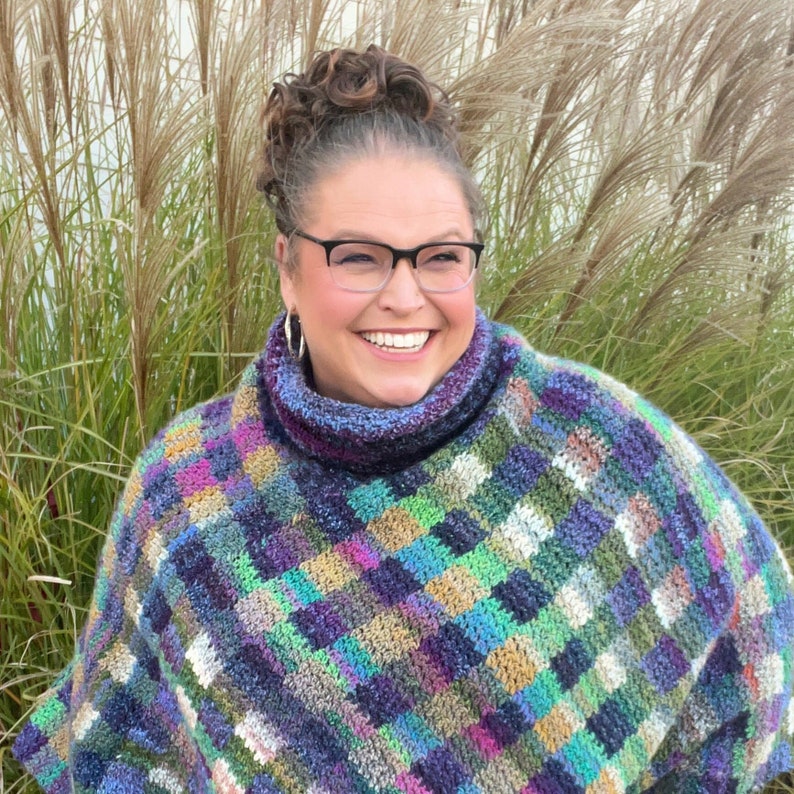 Marly Bird wearing the Check Me Out Gingham Plaid Crochet Poncho. The poncho is a rich tapestry of multicolored squares in hues of purple, green, and blue, with subtle tweed textures. The cowl neckline of the poncho showcases the gingham plaid stitch