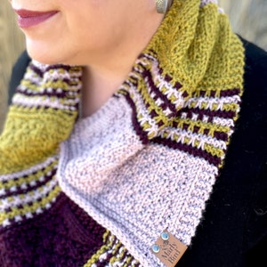 three color knit bandana cowl worn around the neck of Marly Bird. The bind off edge is seamed to the bottom ridge edge near the cast on to form a point in front when wearing it. The cowl is made in light pink, wine purple, and a golden wheat color.