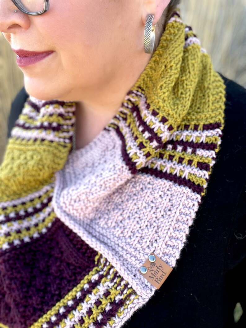 three color knit bandana cowl worn around the neck of Marly Bird. The bind off edge is seamed to the bottom ridge edge near the cast on to form a point in front when wearing it. The cowl is made in light pink, wine purple, and a golden wheat color.