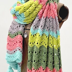 A vividly colored Calor Crochet Wrap by Marly Bird draped over a mannequin, showcasing a wave crochet pattern and bright tassels.