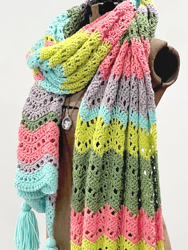 A vividly colored Calor Crochet Wrap by Marly Bird draped over a mannequin, showcasing a wave crochet pattern and bright tassels.