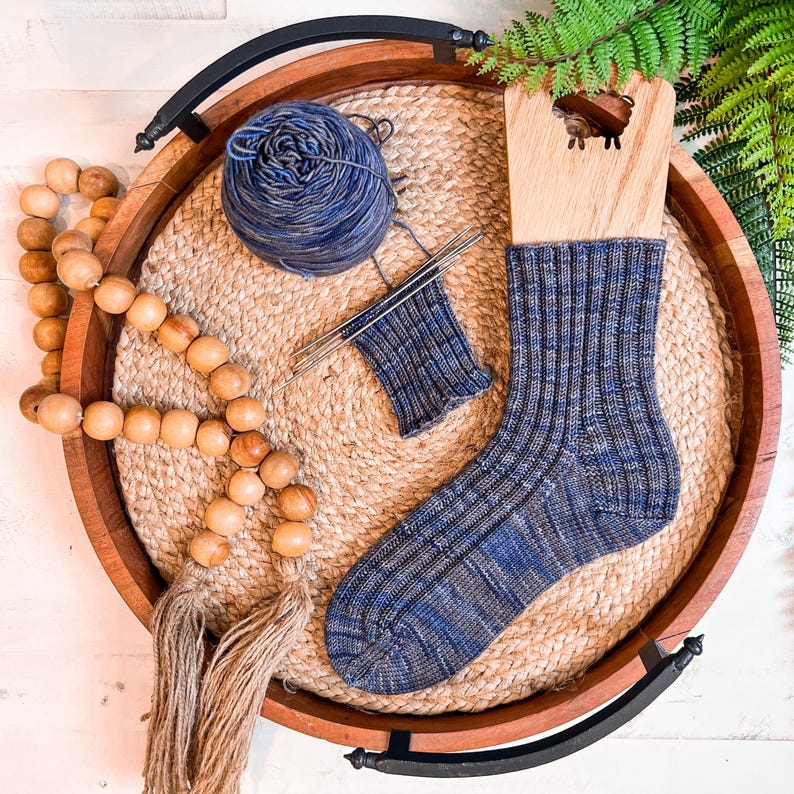 A blue knit sock on a wooden sock blocker, with matching blue yarn and knitting needles, sits on a round woven tray. Wooden beads and green fern leaves decorate this charming scene—perfect as a thoughtful handmade gift.