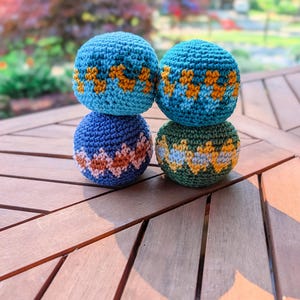 Stack of three crochet hacky sacks in blue and green with zigzag colorwork stripes on a wooden table outdoors.