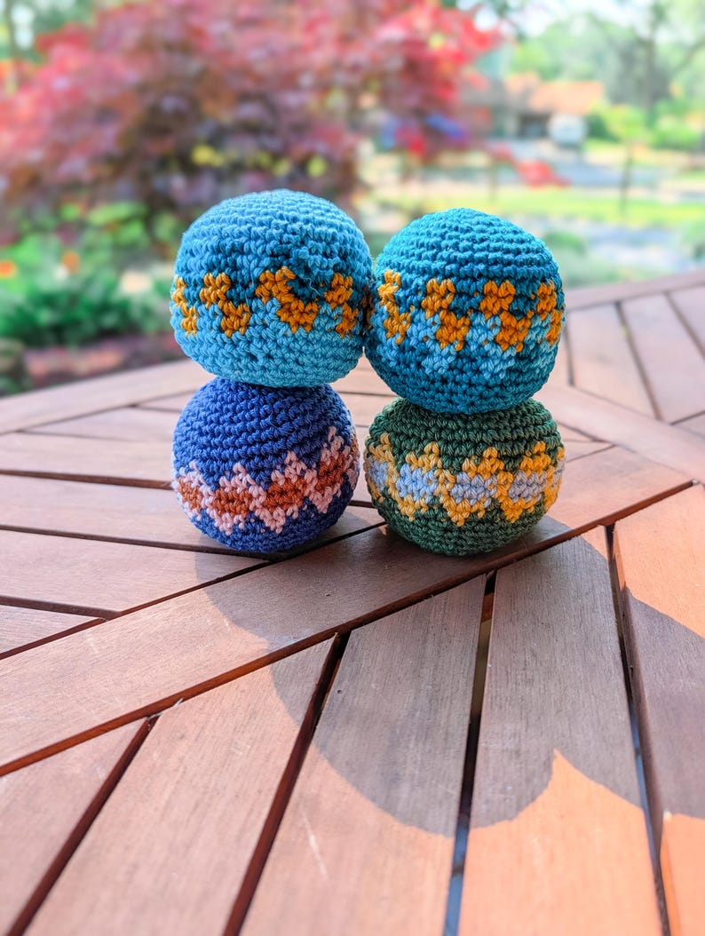 Stack of three crochet hacky sacks in blue and green with zigzag colorwork stripes on a wooden table outdoors.