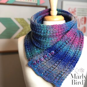 May include: A hand-knitted cowl in shades of blue, purple, and teal, displayed on a mannequin. The cowl features a ribbed texture and is secured with six round, purple buttons. The Marly Bird logo is visible in the lower right corner.