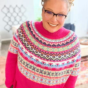 Pode incluir: Um suéter rosa vibrante com gola alta e mangas compridas. O suéter apresenta um design colorido no peito e na parte superior das mangas, incluindo formas geométricas e listras em tons de azul, verde, branco e vermelho. O suéter é feito de malha macia.