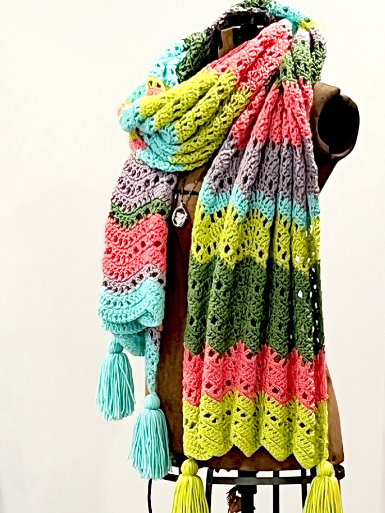 A vividly colored Calor Crochet Wrap by Marly Bird draped over a mannequin, showcasing a wave crochet pattern and bright tassels.