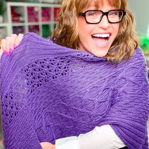 May include: A vibrant purple knitted shawl featuring a complex texture with alternating sections of intricate lace eyelet patterns and raised cable knit designs. The fabric is a deep violet hue, showcasing a detailed stitch work throughout the garment.